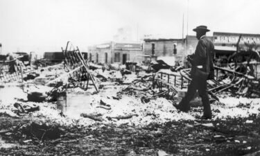 An African American man with a camera looking at the skeletons of iron beds which rise above the ashes of a burned-out block after the Tulsa Race Massacre in Tulsa