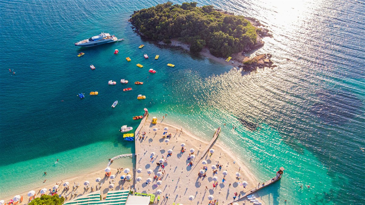<i>MZaitsev/Adobe Stock</i><br/>Aerial view of a beautiful white sand beach with turquoise water and relaxing people on a sunny day. Ksamil