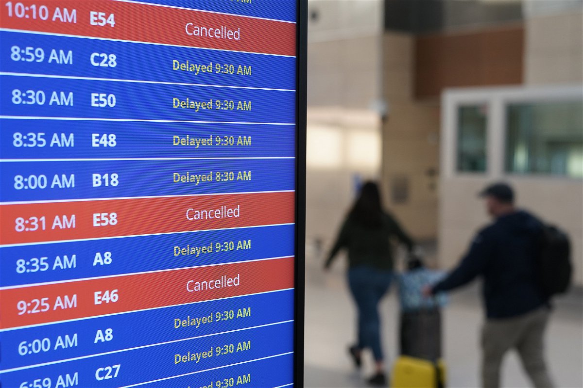 <i>Patrick Semansky/AP</i><br/>Travelers walk as a video board shows flight delays and cancellations at Ronald Reagan Washington National Airport in Arlington