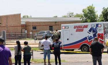 An ambulance waits at Robb Elementary School as people watch from behind police tape in Uvalde