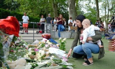 A woman holding a baby lays flowers near the scene at a lakeside park in Annecy