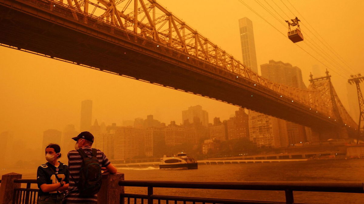 <i>Shannon Stapleton/Reuters</i><br/>People wear protective masks as the Roosevelt Island Tram crosses the East River while haze and smoke from the Canadian wildfires shroud the Manhattan skyline.