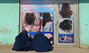 Afghan burqa-clad women sit in front of a beauty salon with images of women defaced using spray paint in Jalalabad on December 13