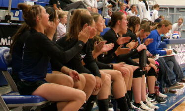 A row of volleyball players in black uniforms sit on a bench and applaud their teammates.